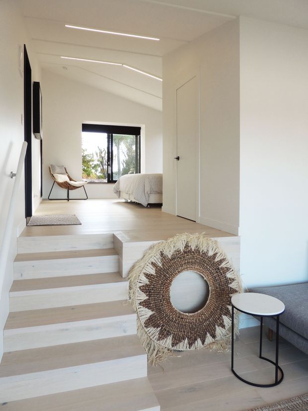 Bright minimalist split-level interior with pale timber flooring and broad white walls. A short flight of light timber stairs leads to a raised bedroom area with a neatly made bed, woven lounge chair, and black-framed window. Linear recessed LED lighting runs across the white ceiling, while a grey sofa and small round side table sit in the foreground beside a large woven circular wall decoration.
