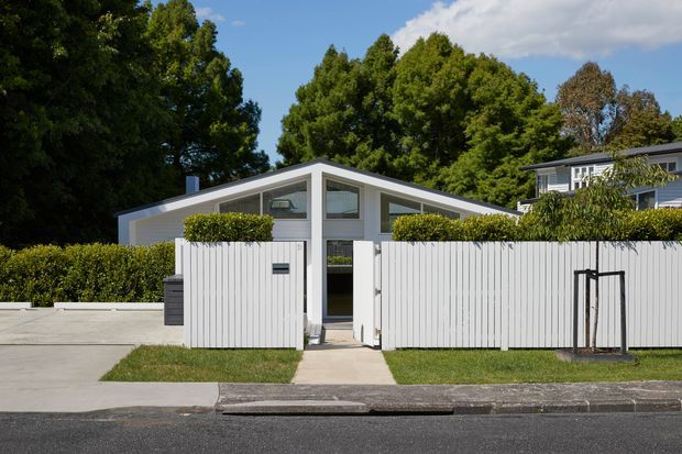 Street-facing view of a low, single-storey residential house behind a white vertical-slat fence and clipped hedges. The home has a distinctive central gable with broad sloping roof planes, pale grey exterior walls, large triangular and rectangular glazing, and a simple pedestrian entry gate. A concrete driveway and footpath connect the property to the street, with mature trees behind the house.