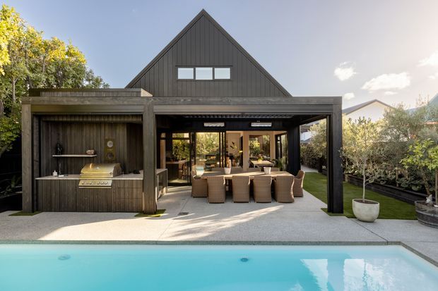 A contemporary dark-clad gabled house viewed from the pool courtyard. A covered outdoor entertaining area contains a dining table and chairs, with a built-in outdoor kitchen or bar to the left. The paved terrace opens directly onto a bright swimming pool, with landscaped planting around the edges.