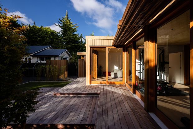 A warm contemporary timber-clad residential extension viewed from a spacious timber deck courtyard. Floor-to-ceiling glazing and timber-framed sliding doors open onto the deck, revealing a light-filled interior with a sofa and built-in elements. The composition also shows a neighbouring gabled home, mature trees, and strong late-afternoon sunlight.