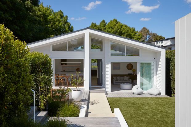 Bright contemporary single-storey pavilion-style home with a distinctive asymmetric pitched roof, extensive white-framed glazing, and a central entrance. The façade opens onto a landscaped courtyard with a timber deck, concrete pathway, manicured lawn, hedges, and ornamental planting. Interior glimpses show a dining area on the left and a relaxed living room on the right.