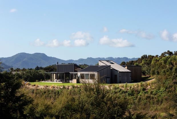 A contemporary single-storey home set within a natural, bushy landscape, with dark vertical cladding, several intersecting pitched roof forms, extensive glazing, and a sheltered terrace. Mountain ranges are visible in the background beneath a clear blue sky.