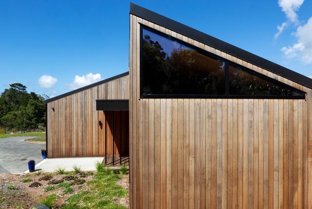 Exterior view of a contemporary single-storey building clad in vertical natural timber boards. The sharply angled roofline and large dark-framed triangular clerestory glazing create a distinctive gabled form, with a recessed timber entry and simple landscaped foreground.