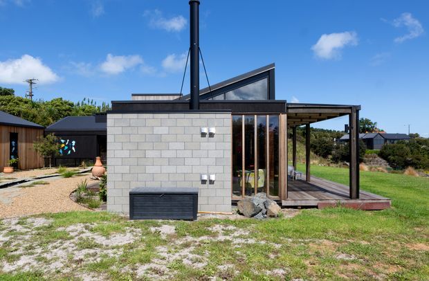 Exterior view of a compact contemporary pavilion-like dwelling set in a landscaped suburban or semi-rural site. The building features a pale grey concrete block wall, dark metal roof and chimney, extensive timber-framed glazing, and an open covered deck or verandah. A sloping clerestory-style roof element rises above the main volume, while gravel paths, planted areas, lawn, and adjacent dark-roofed structures surround the building.