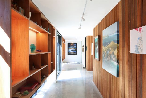 Bright, elongated interior hallway or gallery with polished concrete flooring, extensive warm timber built-in shelving and wall panelling, framed artwork, recessed shelf lighting, and a white ceiling with track lights. Glazed openings and a bright area at the far end create an airy, contemporary gallery-like circulation space.