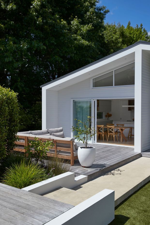 Exterior view of a compact, contemporary white-clad gabled house opening onto a landscaped outdoor entertaining area. The facade has horizontal white cladding, a dark narrow roof edge, high-level glazing, and large sliding doors. A timber deck and paved terrace feature built-in outdoor seating, a white planter, and garden landscaping, while a dining area is visible inside.