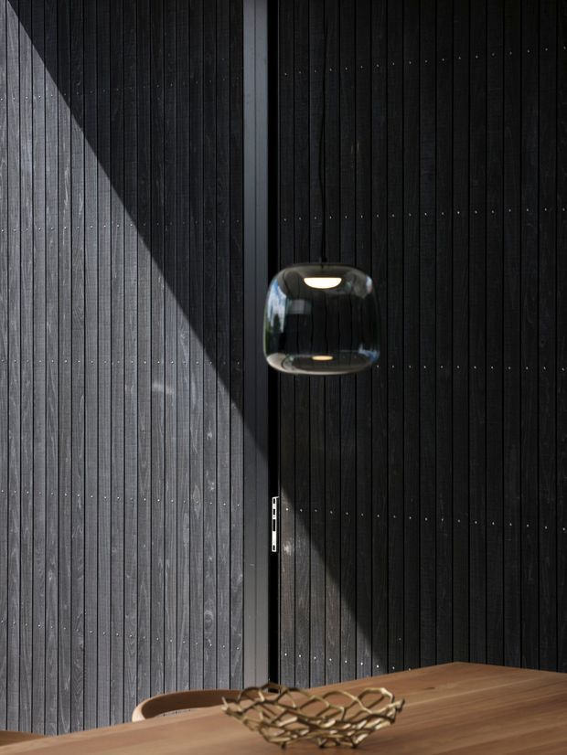 A moody, minimalist interior detail featuring a dark vertical timber-panelled wall and matching timber door, sharply defined by sunlight and shadow. A sculptural black pendant light hangs above a warm timber tabletop with a wire fruit bowl, creating a high-contrast, refined composition.