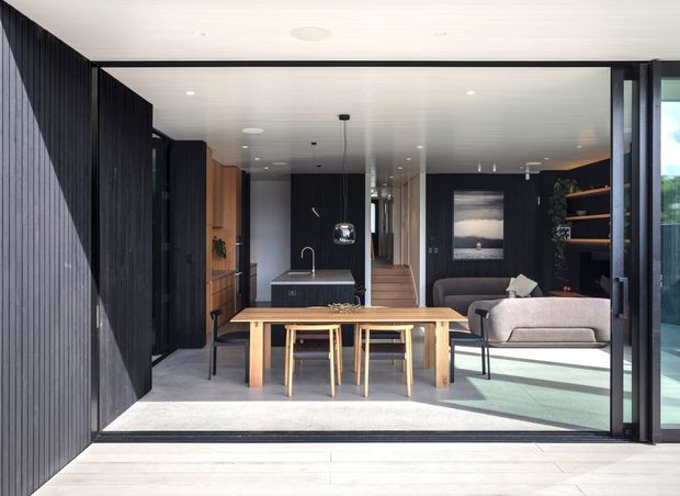 A refined open-plan living and dining space viewed through fully glazed sliding doors. The room features dark vertically ribbed wall panelling and cabinetry, a light timber dining table with matching chairs, a grey sofa, a compact kitchen at the rear, polished concrete flooring, and a bright covered outdoor threshold. The interior has a restrained contemporary character with strong black-and-timber contrast.