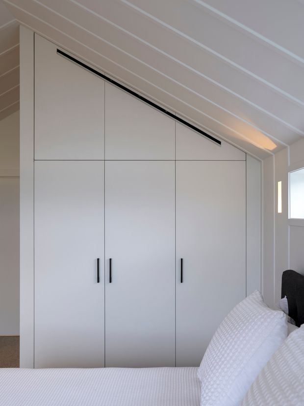 A bright, compact bedroom featuring a full-height built-in wardrobe with clean white panelled doors and slim black handles. The wardrobe follows the sloping ceiling line, incorporating a long black ventilation or lighting slot. White bedding and a dark upholstered bedhead are visible in the foreground. The room has a white painted timber-lined sloped ceiling and small high-set windows.