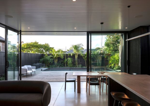 Contemporary open-plan interior with a dining area, kitchen island, and living space opening through extensive floor-to-ceiling glazing onto a timber deck and landscaped pool courtyard. The room has a white linear-panel ceiling, polished concrete flooring, dark vertical wall cladding, warm timber furniture, and a restrained neutral palette.