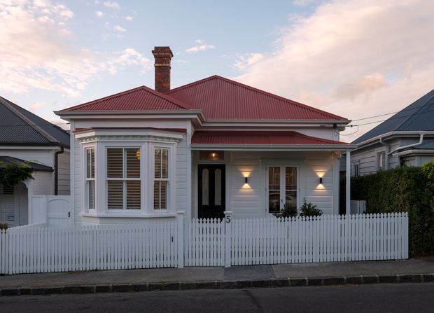 Street-facing view of a compact single-storey heritage-style house with a white weatherboard facade, ornate bay window, front porch, red corrugated pitched roof, brick chimney, and white picket fence. The home sits closely between neighbouring houses in an established suburban streetscape, photographed at dusk with exterior wall lights illuminated.