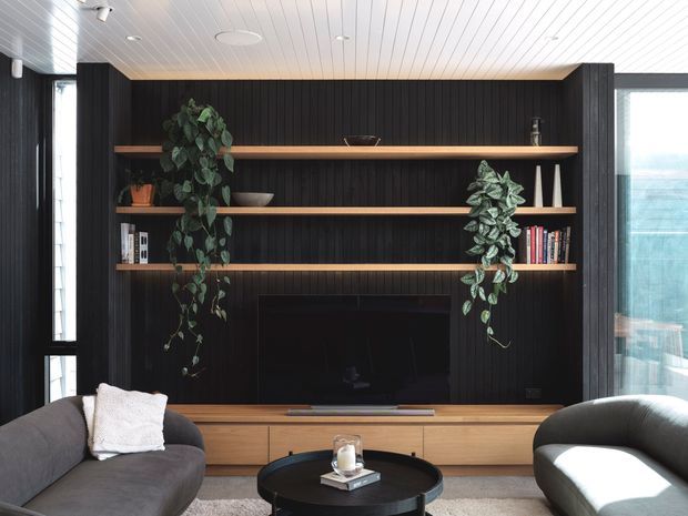 A contemporary living room centered on a built-in entertainment wall with dark vertical timber panelling, open timber display shelves, and a recessed television above low timber cabinetry. Two curved grey sofas frame a round black coffee table, while trailing indoor plants, books, and decorative objects add warmth. A white-lined ceiling and expansive glazing bring in abundant daylight.