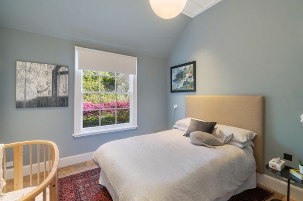 A cozy bedroom with muted blue-grey walls, a white-trimmed window overlooking greenery, a large upholstered bed with neutral bedding, and a wooden cot in the foreground. A patterned rug, framed artwork, bedside table, and warm ceiling light complete the softly styled room.