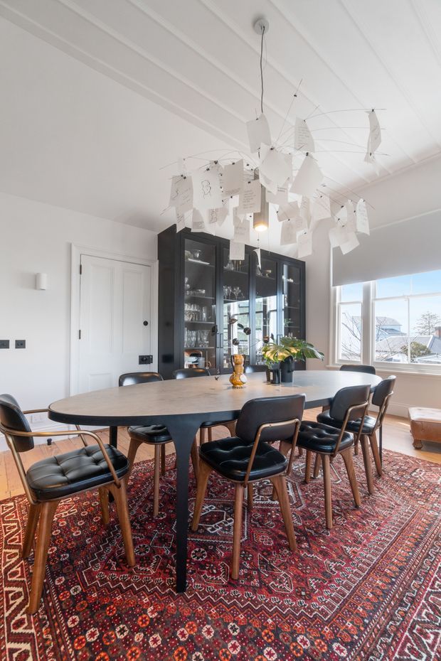 Bright dining room with a large oval timber dining table surrounded by mid-century-style dark leather and timber chairs. A deep charcoal built-in glass-front cabinet displays glassware along the rear wall, while a sculptural hanging installation of white paper forms occupies the coffered white ceiling. A large patterned Persian-style rug grounds the room, and expansive windows provide abundant natural light and views outside.