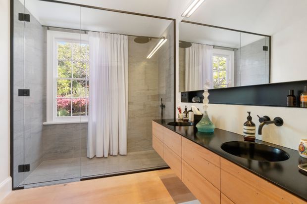 A refined contemporary bathroom with a spacious glass-enclosed walk-in shower, pale large-format wall and floor tiles, white curtains, and a timber floating vanity. The vanity has a dark stone benchtop, integrated black basin, matte black wall-mounted tapware, and a large mirrored wall cabinet. Natural light enters through the window, creating a bright, warm, minimalist atmosphere.