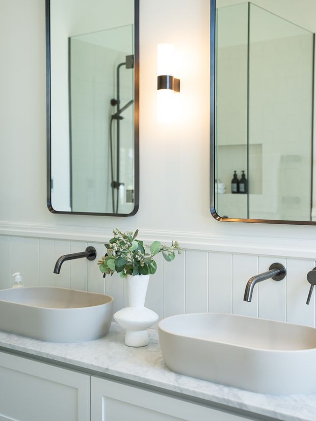 Bright, refined double-vanity bathroom with two white vessel basins on a pale stone countertop, dark wall-mounted tapware, white vertical wall panelling, large rounded-corner mirrors, and a warm wall sconce. A shower is visible in the left mirror reflection, while small decorative greenery adds a natural accent.