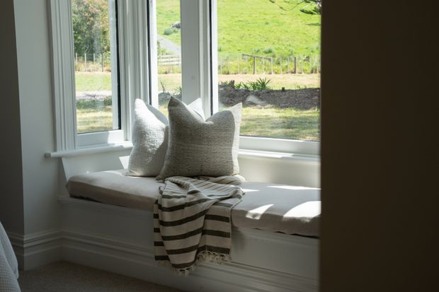 A bright, tranquil window seat set into a projecting bay window, with white-painted trim and built-in white joinery. Neutral textured cushions and a striped throw create a soft, inviting reading nook, while the windows overlook a green landscaped hillside.