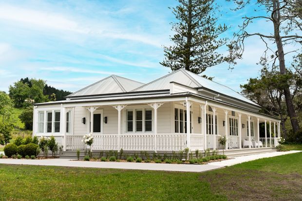 A single-storey cream-white heritage-style house with a broad hipped metal roof, wraparound verandah, decorative timber fretwork, white balustrades, tall shuttered windows, and French doors. The house sits in a landscaped suburban or semi-rural garden with mature trees and lawn under a bright sky.