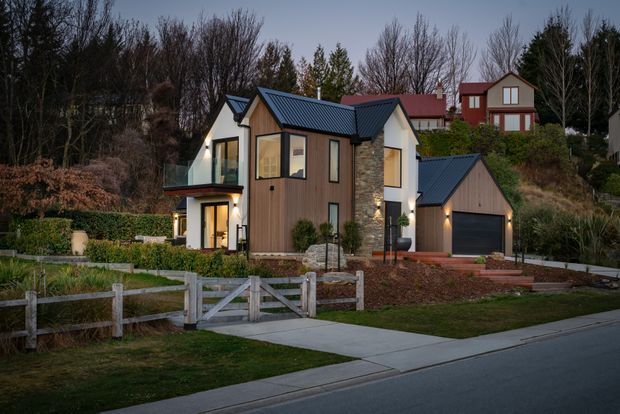 A contemporary two-storey residential home viewed from the street at dusk. The house features dark standing-seam pitched roofs, vertical timber-clad wall sections, pale rendered or light-coloured walls, prominent natural stone cladding, large windows, and a projecting glazed balcony. A detached or connected single-car garage sits to the right, with landscaped gardens, fencing, and a concrete driveway in the foreground. Other houses and mature trees are visible in the background.