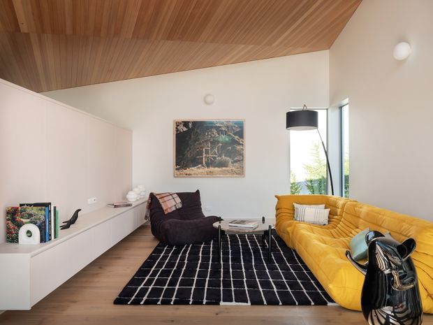 A stylish living room with a warm timber-lined ceiling, white walls, timber flooring, built-in white cabinetry, and large windows. The room features a mustard-yellow curved sofa, dark lounge chair, black-and-white grid rug, sculptural black side table or vase, pendant lighting, and framed artwork.