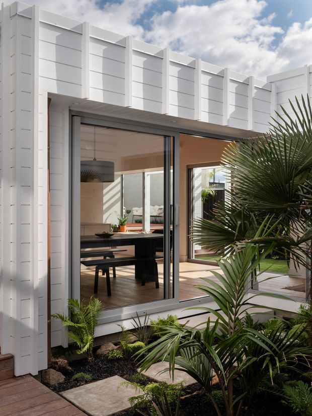 Exterior view of a contemporary white-clad building enclosing a landscaped courtyard. Tall vertical architectural fins and horizontal panel lines create a strong geometric facade, while large glazed sliding doors open to a dining area with a dark table and pendant light. Tropical planting, stepping stones, timber decking, and a warm-toned interior are visible around the courtyard.