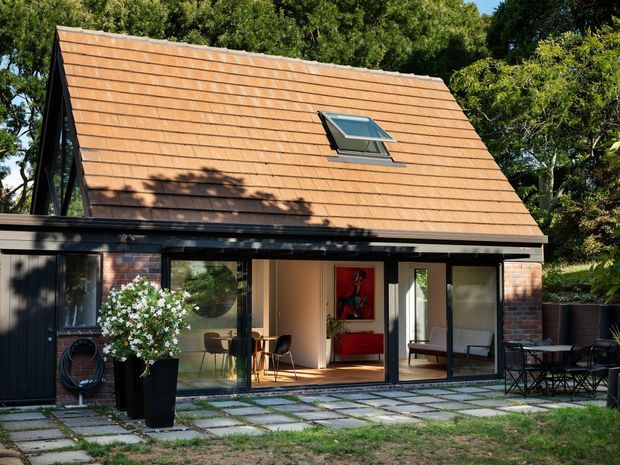 Exterior view of a compact modern gable-roof dwelling set among mature trees. The steep roof is clad in terracotta-toned tiles and includes a roof skylight. Dark vertical cladding, exposed red brick sections, extensive black-framed glazing, and large sliding doors open onto a paved patio with outdoor dining furniture. Interior living spaces are visible through the glazing.