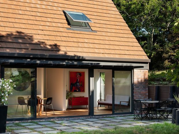A compact modern extension or pavilion with a steeply pitched terracotta tile roof, roof skylight, dark-framed floor-to-ceiling glazing, and exposed brick side wall. The open glazed facade reveals a bright interior with a dining area and red built-in cabinetry. A paved terrace with outdoor dining furniture occupies the foreground, surrounded by mature garden planting.