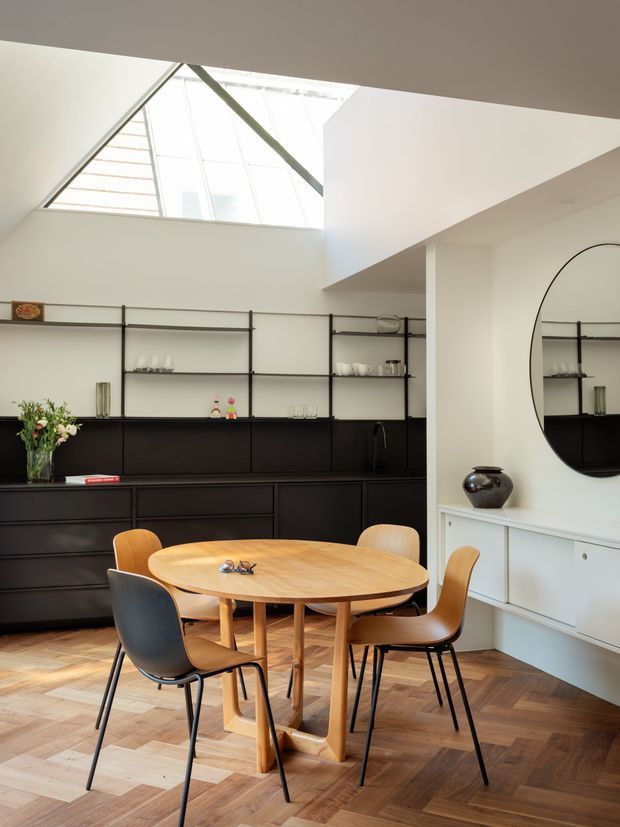 A bright contemporary dining area with a round light-wood dining table and mixed wooden and dark upholstered chairs. A full wall of black built-in cabinetry with open black metal shelving provides storage and display space. The room features a geometric parquet timber floor, white walls, a large round wall mirror, and a dramatic angular ceiling opening with a skylight that brings in abundant natural light.
