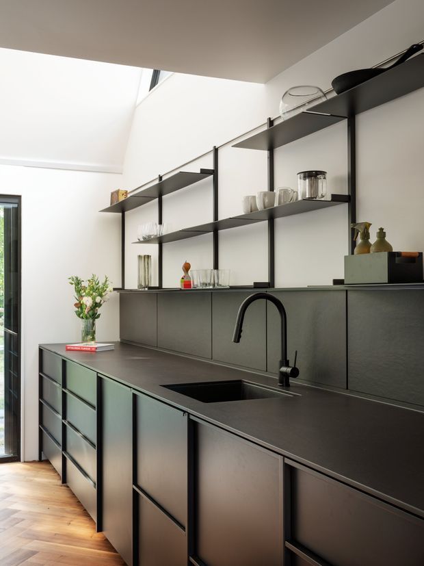 A contemporary kitchen with dark matte cabinetry, a large island with integrated black sink and arched faucet, and open black metal shelving displaying glassware and ceramics. White walls and a high ceiling with a skylight or clerestory opening create contrast, while timber flooring adds warmth. A glazed door or window is visible at the left.