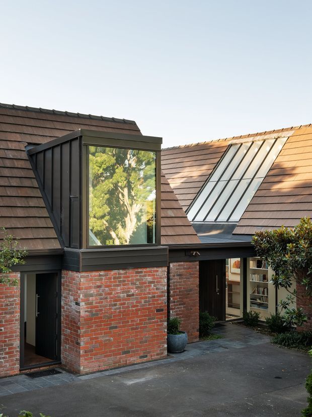 Exterior view of a compact residential building arranged around a paved courtyard. The structure features red brick walls, dark-framed projecting glazing, steep shingle-clad pitched roofs, and a prominent slanted roof skylight. A partially open doorway reveals shelving and an interior passage, while planting borders the courtyard.