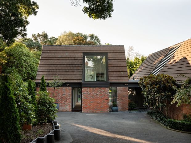 Exterior view of a compact contemporary brick house set within a mature landscaped garden. The building features a steeply pitched dark shingle-clad upper volume, exposed red brick walls, dark-framed glazing, and a low connecting roof or entry canopy. A paved driveway or forecourt leads toward the recessed entrance, with a neighbouring pitched-roof structure visible at right.