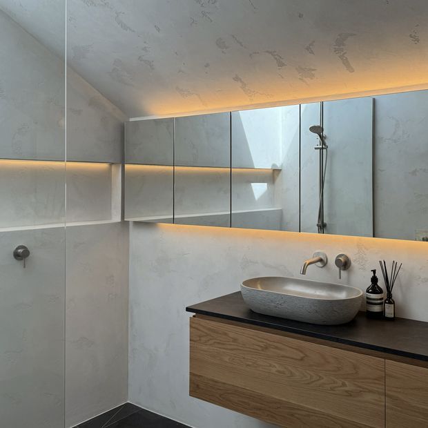 Contemporary bathroom featuring pale textured plaster or stone-look wall and ceiling surfaces, a floating natural timber vanity with a dark benchtop, a sculptural vessel basin, brushed metal tapware, mirrored wall cabinetry, integrated warm LED lighting, and a frameless shower area.