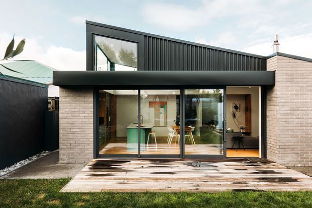 Exterior view of a compact contemporary single-storey extension or dwelling with a dark vertically ribbed metal upper facade, pale brick side walls, large black-framed glazed sliding doors, and a timber deck leading into an open-plan interior. The composition shows a flat-roofed form with strong horizontal and vertical lines.