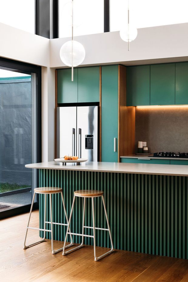 A bright contemporary kitchen with a tall double-height ceiling and clerestory glazing. Teal-green flat-panel cabinetry is framed by warm timber panels, with a dark textured backsplash, integrated stainless-steel refrigerator, gas cooktop, and a large pale island bench. The island has a vertically fluted green base and two timber-and-metal stools. Globe pendant lights hang above, while warm timber flooring and extensive black-framed glazing add contrast.