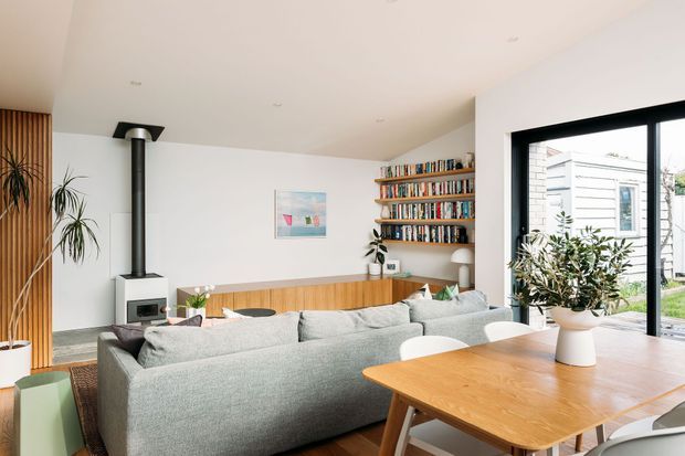 Bright open-plan living and dining area with a large light-grey sectional sofa, timber dining table, built-in timber wall panelling, floating bookshelves, a freestanding wood-burning stove, indoor plants, and large black-framed glazing opening toward a garden. The room has a clean, contemporary aesthetic with warm natural timber accents.
