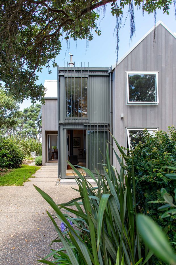 Exterior view of a contemporary multi-level dwelling set within a leafy landscaped garden. The facade combines dark grey vertical cladding with a steep gabled volume, flat-roofed sections, large glazed openings, and a tall screened window or balcony enclosure. A concrete path leads toward an open entry, with dense ornamental planting framing the approach.