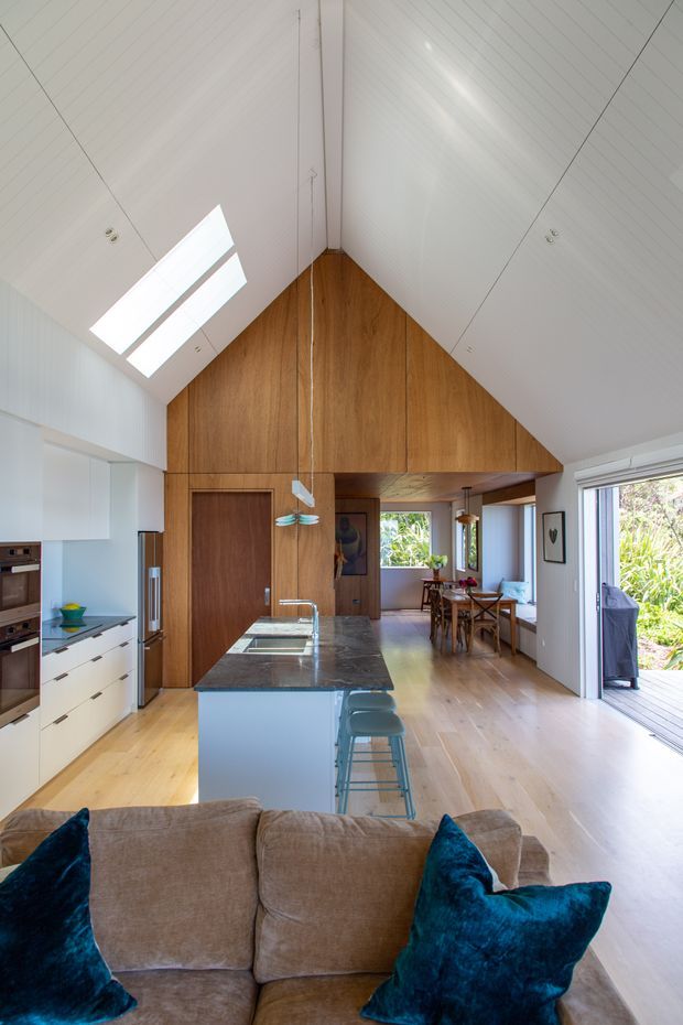 A bright, open-plan kitchen and living area with a dramatic vaulted ceiling. The kitchen features white fixed cabinetry, integrated appliances, a dark stone island benchtop, timber flooring, and a warm timber-clad gable wall with a concealed door. Bar stools line the island, while a dining area and living room with a large sofa are visible beyond. Skylights, clerestory-style glazing, and wide glazed doors provide abundant natural light and connect the interior to an outdoor deck.