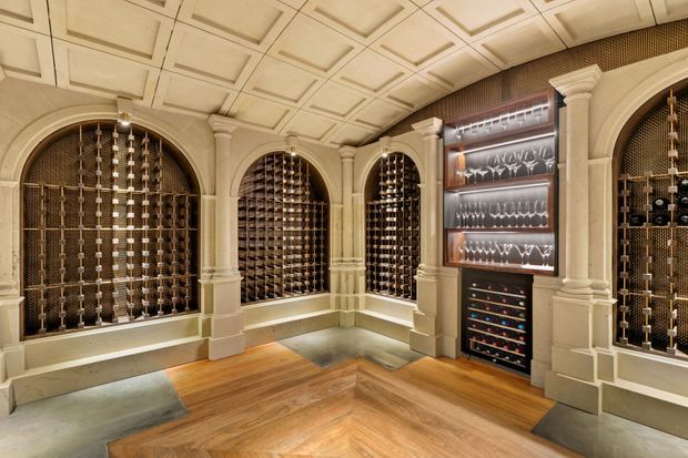 A luxurious wine cellar with multiple arched, column-framed wine storage niches, extensive bottle racks, and a central glassware and wine display cabinet. The room features ornate coffered ceiling panels, classical architectural detailing, stone or painted masonry surrounds, and a warm chevron-pattern timber floor.