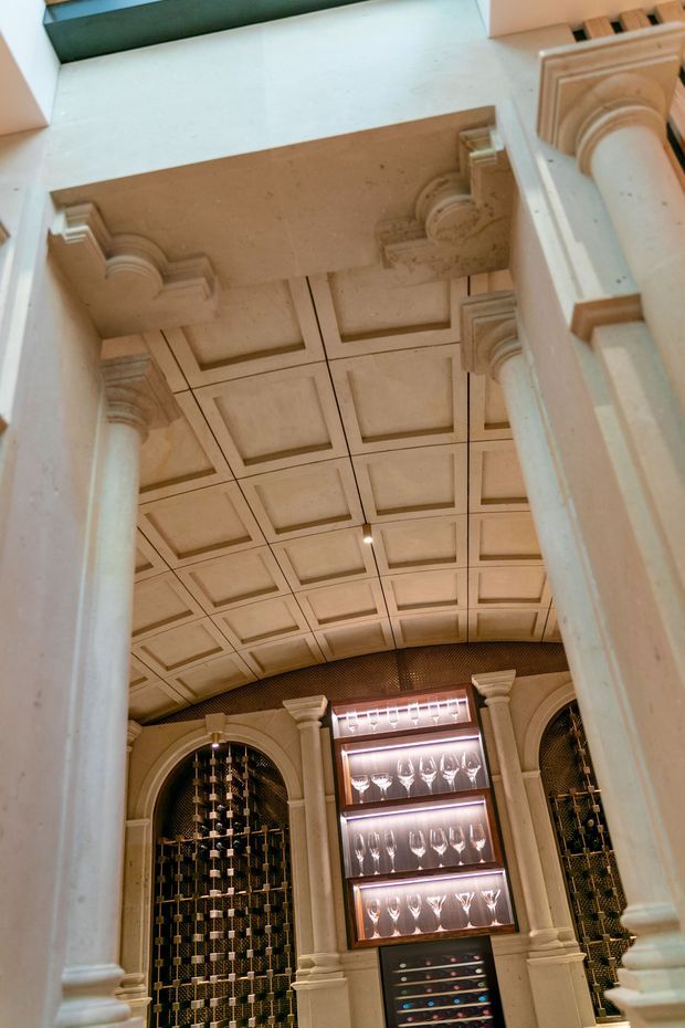 Interior view of an ornate wine cellar or wine room framed by classical columns and decorative arches. The room features an elaborate coffered ceiling, carved architectural mouldings, dark wine bottle storage, and illuminated glassware display cabinetry.