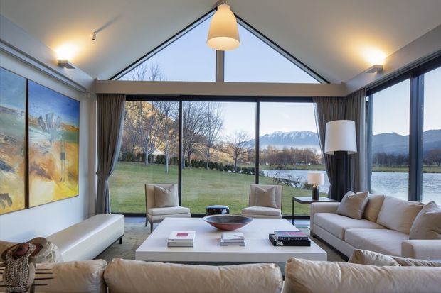 A bright, contemporary living room with expansive floor-to-ceiling glazing overlooking a lawn, lake, and mountain landscape. The room features a large cream sectional sofa, two upholstered lounge chairs, a white rectangular coffee table, floor and pendant lighting, curtains, and a colourful wall artwork. A dramatic triangular clerestory window and exposed central structural post define the ceiling composition.