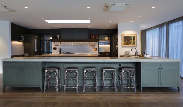 A spacious contemporary kitchen centred on a long blue-grey island with integrated cabinetry and five metal bar stools. The island faces a dark built-in kitchen wall with illuminated glass-front display cabinets, a tiled or stone backsplash, cooktop and concealed appliances. A white benchtop continues around the room, with a sink and tap beneath large sheer-curtained windows. Warm timber flooring, recessed ceiling lighting, and a framed artwork create a refined, inviting atmosphere.