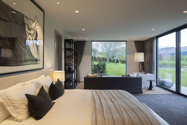 A refined bedroom with a large bed in the foreground, white bedding, dark accent cushions, and a textured neutral throw. The room opens into a bright sitting area with a sofa, side tables, shelving, and floor lamps. Large windows and glazed doors provide expansive views of landscaped grounds, a lake, and distant hills. The interior has a restrained neutral palette, dark curtains, recessed ceiling lights, and contemporary detailing.