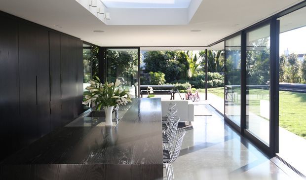 A bright contemporary open-plan kitchen and dining space with a long dark built-in kitchen wall and island, polished dark benchtops, woven dining chairs, and extensive floor-to-ceiling glazing opening toward a landscaped garden. A large roof skylight brings additional daylight into the room.