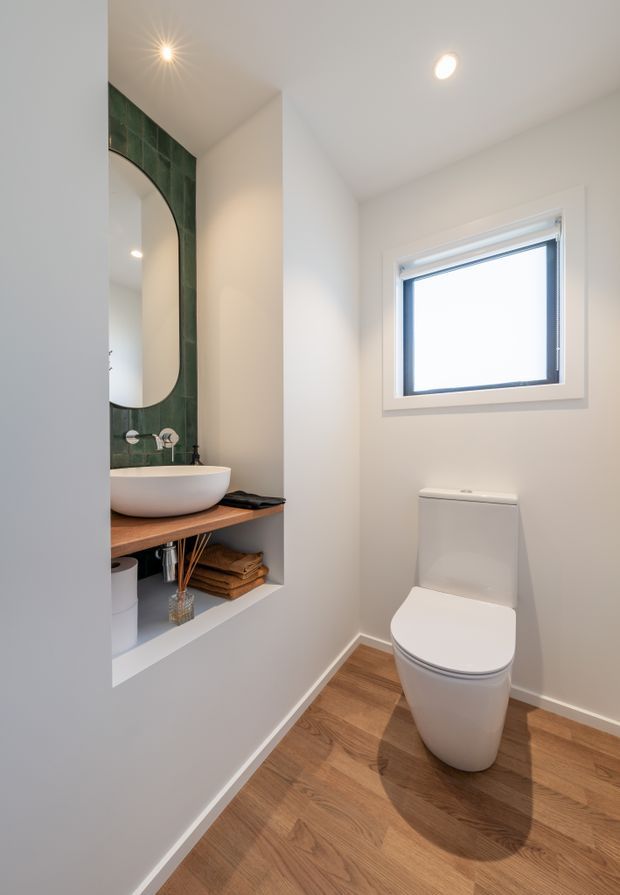 A compact contemporary bathroom featuring a white toilet, wall-mounted basin on a timber vanity shelf, green tiled feature wall with a rounded mirror, recessed storage, white walls, and warm timber-look flooring. A small frosted or bright window provides natural light.
