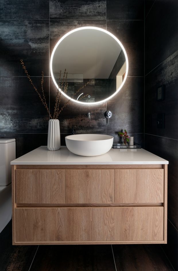 A compact, refined bathroom featuring a floating light-wood vanity with integrated drawers, a white vessel basin, and a white stone-look benchtop. Large dark textured wall and floor tiles create a dramatic backdrop, complemented by a circular illuminated mirror, chrome wall-mounted tapware, and understated decorative styling.