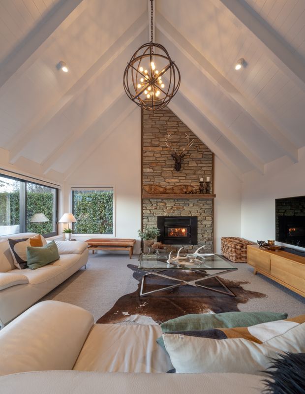 A warm, spacious living room with a dramatic vaulted, white-painted timber ceiling and exposed beams. A central stone-clad fireplace rises to the gable, complemented by a sculptural spherical pendant light. The room includes large windows, cream sectional seating, a glass coffee table, timber media cabinetry, woven baskets, and a mounted television. Natural textures and muted earthy tones create a comfortable lodge-inspired atmosphere.