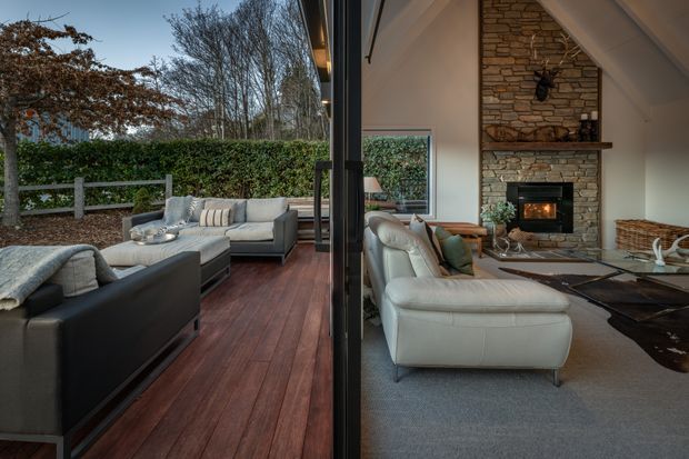 A split indoor-outdoor living space viewed through fully opened dark-framed glazing. The exterior features a timber deck with light upholstered outdoor seating, a hedge-lined garden, and a privacy fence. Inside is a warm, spacious living room with pale sofas, a large stone fireplace surround, timber mantel, wood-burning fire, neutral carpet, and a dark organic-shaped rug. The interior and exterior are visually connected, with a calm, refined residential feel.