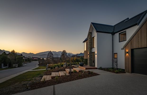 Exterior architectural photograph at dusk showing a contemporary multi-wing house on a landscaped sloping site. The home combines white rendered walls, dark pitched metal roofs, natural stone detailing, and vertical timber cladding around a black garage door. A paved driveway and stepped front path lead through low planting, with mountain ridges visible in the distance.