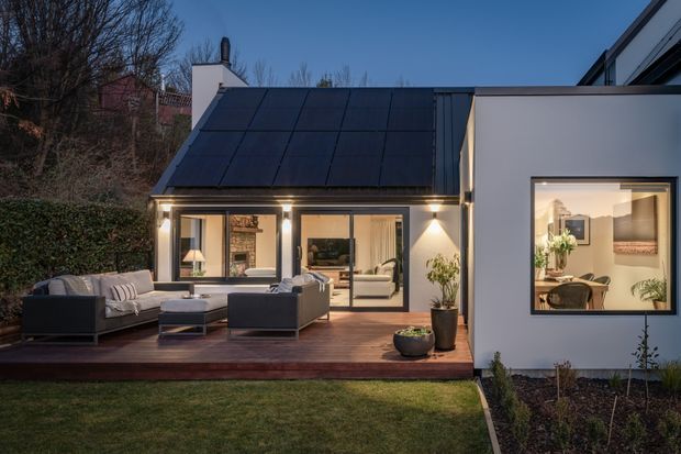 Exterior twilight view of a contemporary single-storey home with a steep dark metal roof, white rendered walls, and large glazed openings connecting the interior living spaces to a timber outdoor deck. The deck includes outdoor lounge seating and potted plants, while warm interior lighting reveals a living room and dining area through the glazing. A landscaped lawn and garden border the house.