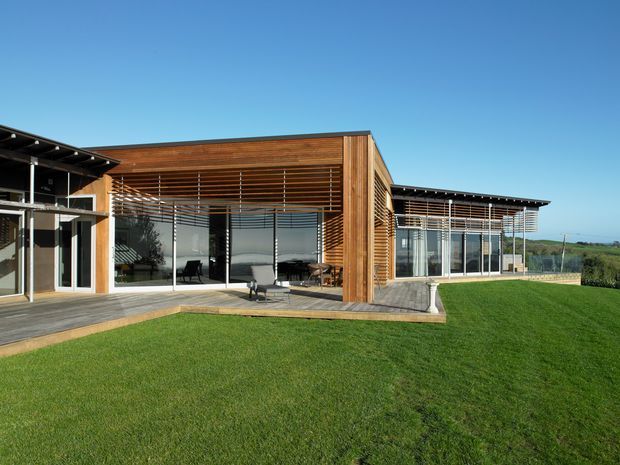 Exterior view of a contemporary single-storey house with extensive floor-to-ceiling glazing, warm timber screens and cladding, broad low-pitched roof planes, and a timber deck opening onto a manicured lawn. The design forms a sheltered courtyard-like outdoor space and overlooks an open rural or coastal landscape.